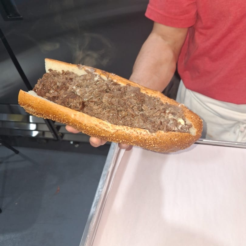 A person in a red shirt holds a steaming, long cheesesteak sandwich with a sesame seed bun.
