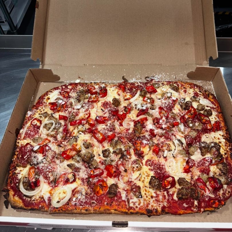 A rectangular Malatesta's pizza with sausage, red peppers, onions, and cheese in a cardboard box.