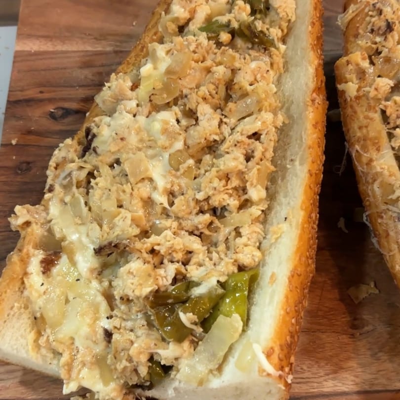 Two halves of a Malatesta's chicken cheesesteak sandwich with onions and green peppers on a wooden board.