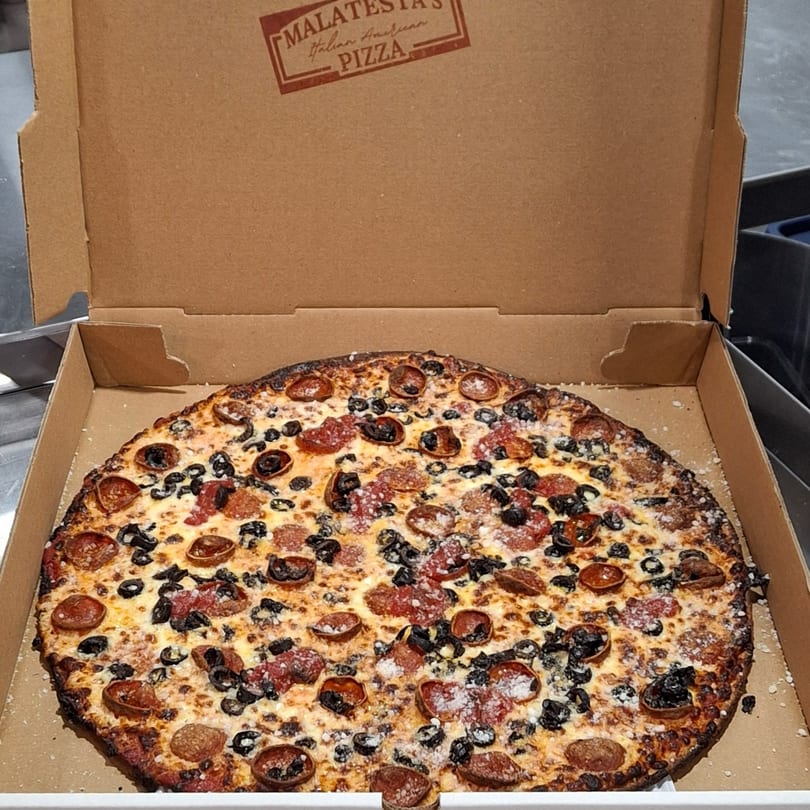 A Malatesta's pepperoni and black olive pizza, sprinkled with parmesan, in a cardboard box.